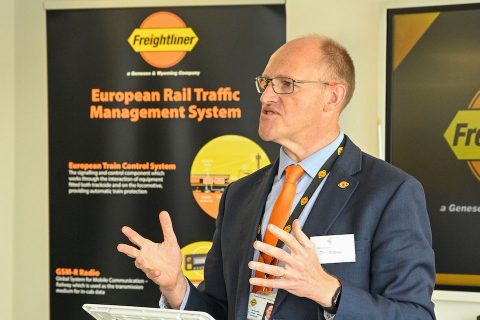 Candid shot of Freightliner CEO Tim Shoveller gesturing with hands while delivering a lecture. Freightliner logo in background